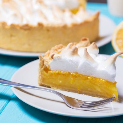 A Slice of Vegan Lemon Meringue Pie Sits on a White Plate With the Pie in the Background, All on a Sky Blue Wooden Table.