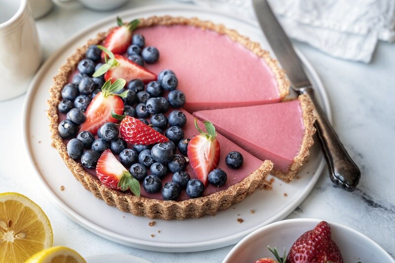 A Tart-Style Pink Lemonade Pie is Decorated With Blueberries and Strawberries With One Slice Ready to Serve.