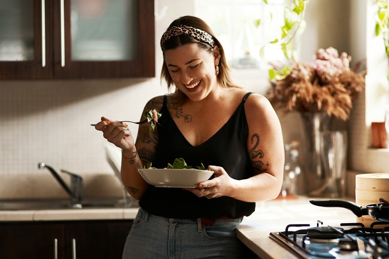 A Woman Smiles as she Eats a Veggie-Filled Meal in a Sun-Lit Kitchen.