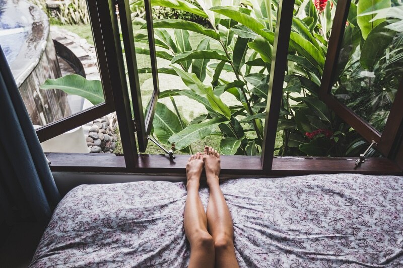 Bedroom Windows are Open, Revealing Lush Plants and a Woman Reclines on a Bed, Representing How to Sleep in the Heat.