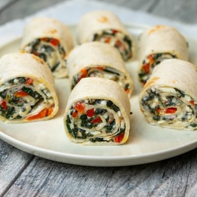 A Round While Plate Has Six Vegan Pinwheel Sandwiches, Beautifully Displaying the Filling With Red Pepper, Spinach and Vegan Cheeses.