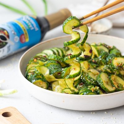 A Rimmed White Plate is Filled With Spiraled Spiced Cucumber Salad Next to a Bottle of Coconut Secret Coconut Aminos.