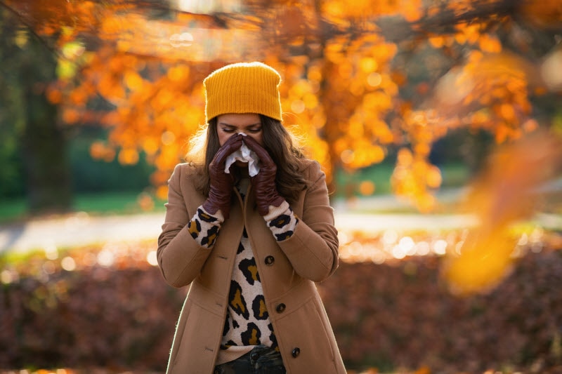 Woman in Coat and Hat Blowing Nose to Represent Fall Allergies Concept