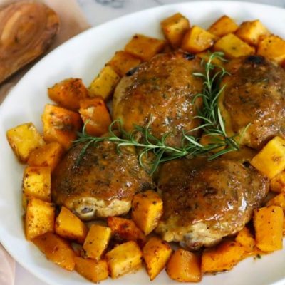 Roasted Chicken Thighs are Nestled in Cubes of Butternut Squash on an Oval Serving Platter Garnished with Sprigs of Rosemary