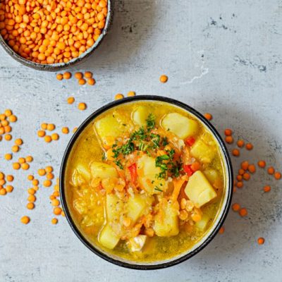 Vegan Lentil Soup with Potatoes in Bowl on Grey Counter Surrounded by a Scattering of Red Lentils