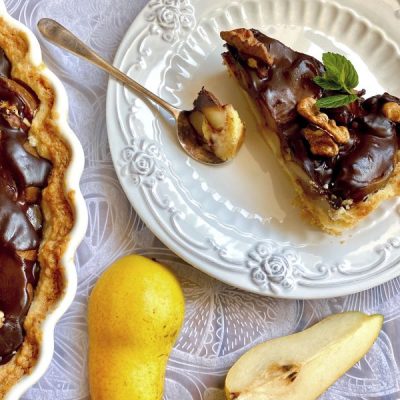 Slice of Chocolate Pear Tart Topped with Walnuts and Mint on Fancy Plate Next to Tart Pan with Whole Tart