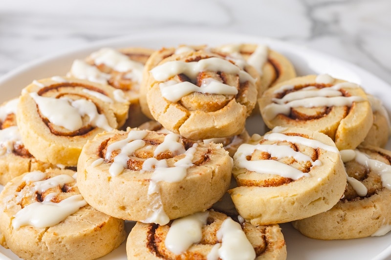 A Plate of Cinnamon Roll Cookies, Each With a Drizzle of Cream Cheese Frosting, Sit Piles on a White Plate.