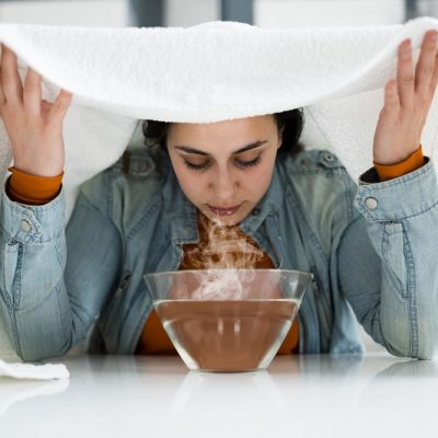 A Woman Uses Essential Oils for Sore Throat Relief, Draping a Towel Over Her Head and Trapping Steam from a Bowl of Hot Water.
