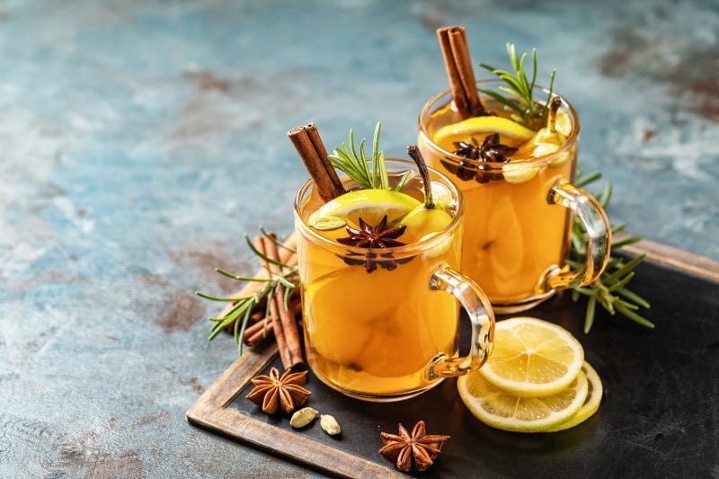 Two Mugs of Holiday Tea with Cinnamon Sticks and Lemon Slices on Chalkboard Atop Blue Marbled Surface