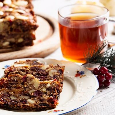Rum-Infused Fruit Cake Topped With Cranberries and Pecans on Fancy Christmas China Next to Hot Tea in a Clear Glass Mug