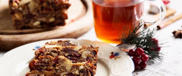 Rum-Infused Fruit Cake Topped With Cranberries and Pecans on Fancy Christmas China Next to Hot Tea in a Clear Glass Mug