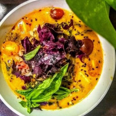 An Overhead Photograph Shows a Bowl of Thai Curry Noodles in a Coconut-Peanut Broth With Fresh Purple Cabbage, Sliced Cherry Tomatoes and Fresh Basil.