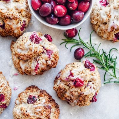 An Overhead View Shows Vegan Drop Biscuits With Cranberries, a Drizzle of Icing, a Small Bowl of Fresh Cranberries and a Sprig of Rosemary.