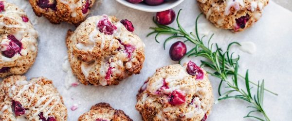 An Overhead View Shows Vegan Drop Biscuits With Cranberries, a Drizzle of Icing, a Small Bowl of Fresh Cranberries and a Sprig of Rosemary.