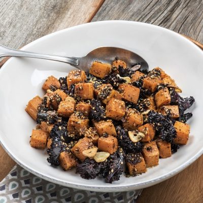 A Warm Kale Salad With Cubed, Roasted Sweet Potatoes and Garlic is in a White, Wide Serving Bowl.
