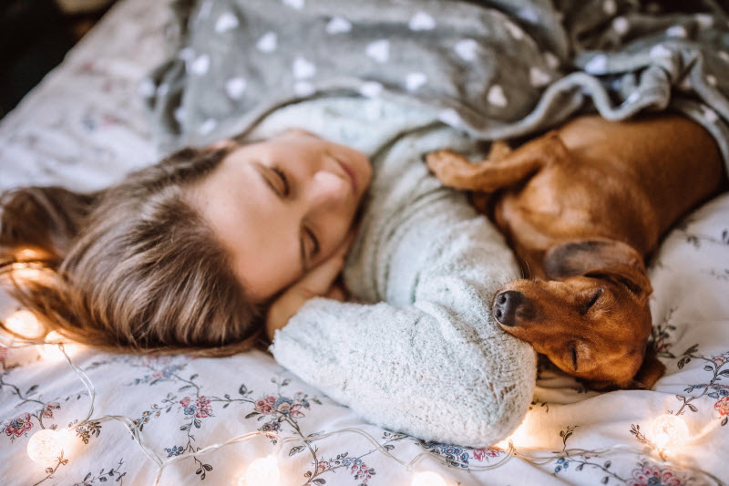 Woman Following Holiday Sleep Tips Cozied up in Bed With Her Dog