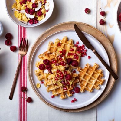 Gluten-Free Waffles on Plate. Sprinkled on Top are Pomegranate Arils and Raspberries along with Granola. There is a Festive Feeling to the Table.