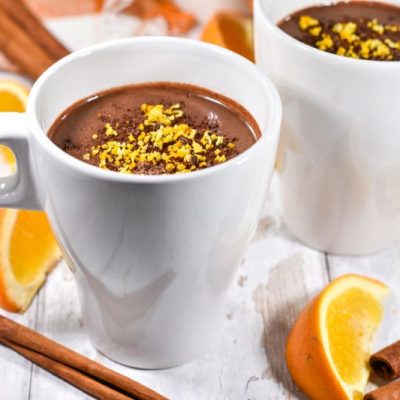 Two Mugs of Orange Hot Chocolate Surrounded by Cinnamon Sticks and Orange Slices on White-Washed Wooden Table