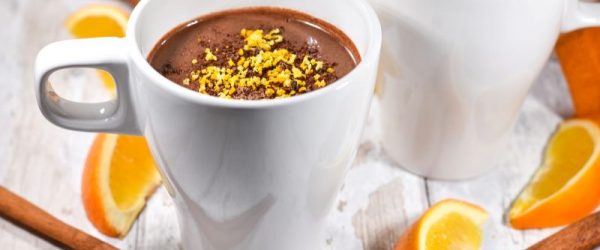 Two Mugs of Orange Hot Chocolate Surrounded by Cinnamon Sticks and Orange Slices on White-Washed Wooden Table