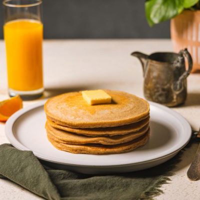 Gluten-Free Buckwheat Pancake Recipe: Pancakes on Plate Topped with a Pat of Butter, a Syrup Pourer Nearby, as well as a Glass of Fresh Orange Juice