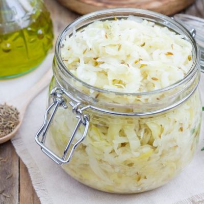 Sauerkraut Recipe Shown in Glass Jar With Lid On a White Cloth Atop a Wooden Table Next to a Spoonful of Caraway Seeds