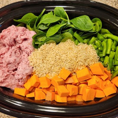 An Overhead View of a Slow Cooker Pot Shows Sections of Pink Ground Meat, Fresh Spinach, Green Beans, Cubed Sweet Potatoes and Dry Brown Rice, Representing How to Make Homemade Dog Food.