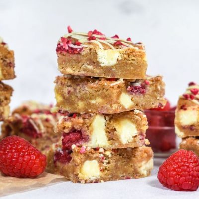 Raspberry White Chocolate Blondies Stacked on Table With Whole Raspberries Scattered Around