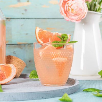 Red Grapefruit Mocktail with Mint Leaves in a Glass Next to a Glass Pitcher set Against a Blue-Painted Wooden Background