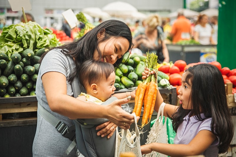 A Mother in the Produce Section of a Grocery Store With Her Two Young Children Buying Carrots, Representing a Plant Based Diet on a Budget.