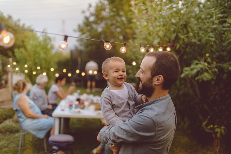 A Father Carries His Toddler Son, and They Smile Together Amongst a Backyard Gathering, Representing Spring Traditions.