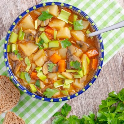 An Overhead View of a Blue Bowl Filled to the Rim With Vegan Irish Stew on a White and Green Checkered Cloth Napkin With Sliced Soda Bread Served on the Side.