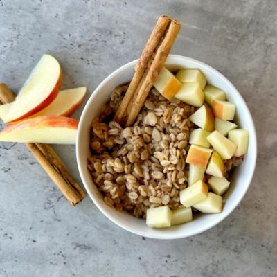 Apple-Cinnamon Barley Porridge in White Bowl on Slate Countertop with Cinnamon Stick and Apple Pieces