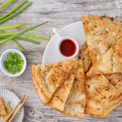 An Overhead View of a Plate of Golden Brown Scallion Pancakes Surrounded by Fresh Scallions and a Dipping Sauce.