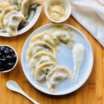 An Overhead View Shows a White Plate Covered in Blueberry Pierogi With a Helping of Greek Yogurt Sauce.