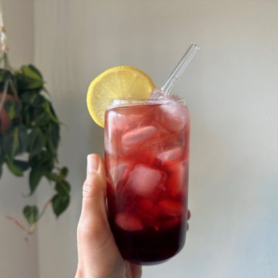 Citrus-Infused Sleepy Girl Mocktail Recipe in Tall Clear Glass With Ice & Straw Held Aloft Next to Hanging Plant in Bedroom