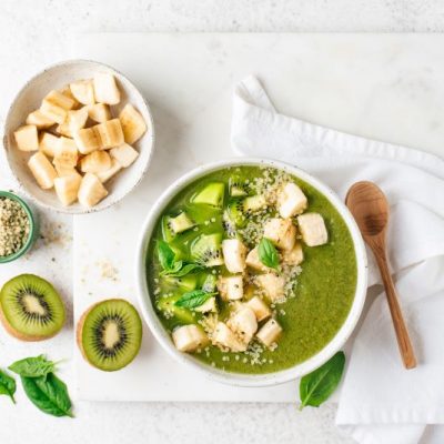 Creamy Green Smoothie Bowl Topped with Slices of Banana and Kiwi on White Marble Counter