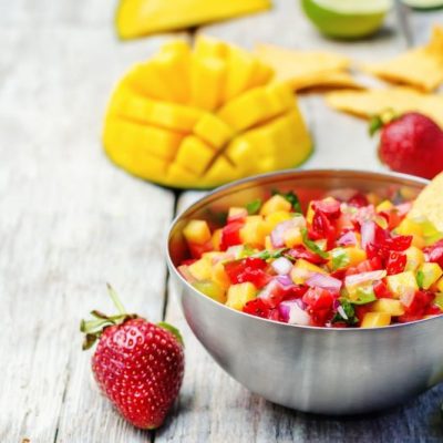 Strawberry Salsa in a Metal Bowl on Tabletop Strewn with Tortilla Chips, Strawberries, Mangoes & Limes