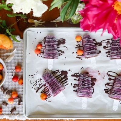 Overhead View of Chocolate-Cherry Ice Pops on Parchment-Lined Sheet Pan Covered in Wild Drizzles of Chocolate, Whole Cherries and Beautiful Flowers Surround the Tabletop Scene