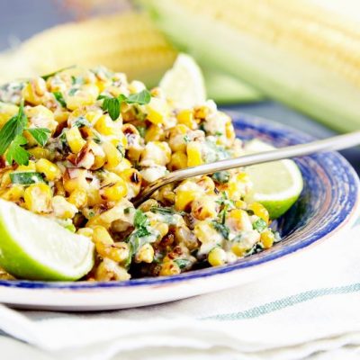 Close up of homemade vegetarian Mexican corn salad with black beans, cilantro and lime, on blue plate on stone background with two ears of corn on blurred background
