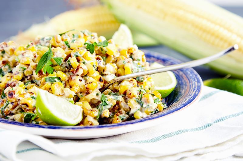 Close up of homemade vegetarian Mexican corn salad with black beans, cilantro and lime, on blue plate on stone background with two ears of corn on blurred background