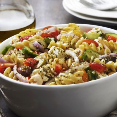 Gluten-Free Greek Pasta Salad in White Bowl Next to Rustic Silver Serving Utensils and a Bottle of Olive Oil