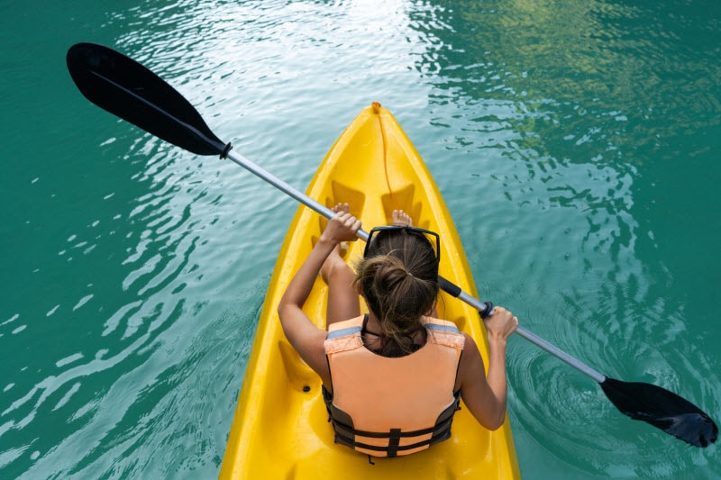 Concept of Is Kayaking Good Exercise Represented by Back-Angle Overhead View of Woman in Yellow Kayak Paddling Along Lake