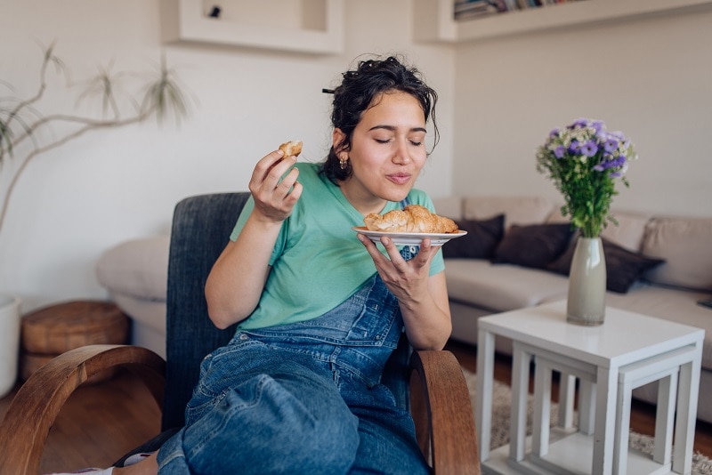 A Young Woman Closes Her Eyes and Savors a Croissant, Representing the Anti Diet Movement.