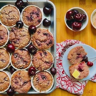 An Overhead View Shows a Dozen Vegan Cherry Almond Muffins With Fresh Cherries Scattered Around.