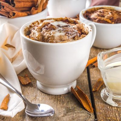 A Cinnamon Bun Mug Cake Sits on a Wooden Table Beside a Cloth Napkin, Cinnamon Sticks and a Small Glass Container of Icing.
