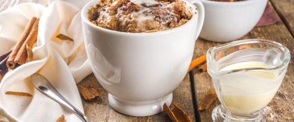 A Cinnamon Bun Mug Cake Sits on a Wooden Table Beside a Cloth Napkin, Cinnamon Sticks and a Small Glass Container of Icing.