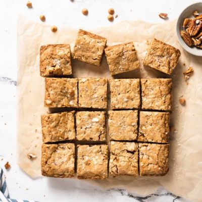 Gluten-Free Butterscotch Blondies on Parchment Paper on Top of Marble Conter Next to a Linen Kitchen Towel and a Small Bowl of Pecans
