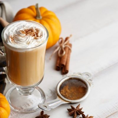 Pumpkin Spice Latte in Footed Glass Mug with Dairy-Free Whipped Cream on Top, Set Next to Miniature Pumpkins, Cinnamon Sticks and Star Anise on Rustic White-Painted Tabletop