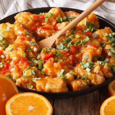 Orange Chicken In Round Black Bowl Surrounded by Slices of Orange on Wooden Countertop