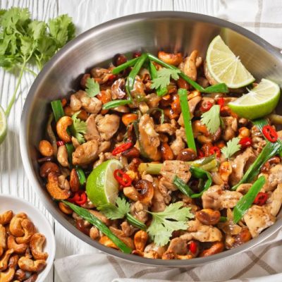 Thai Cashew Chicken in a Metal Serving Bowl Topped With Limes and Cilantro, set Next to a Bowl of Toasted Cashews on Which Rests a Pair of Bamboo Chopsticks